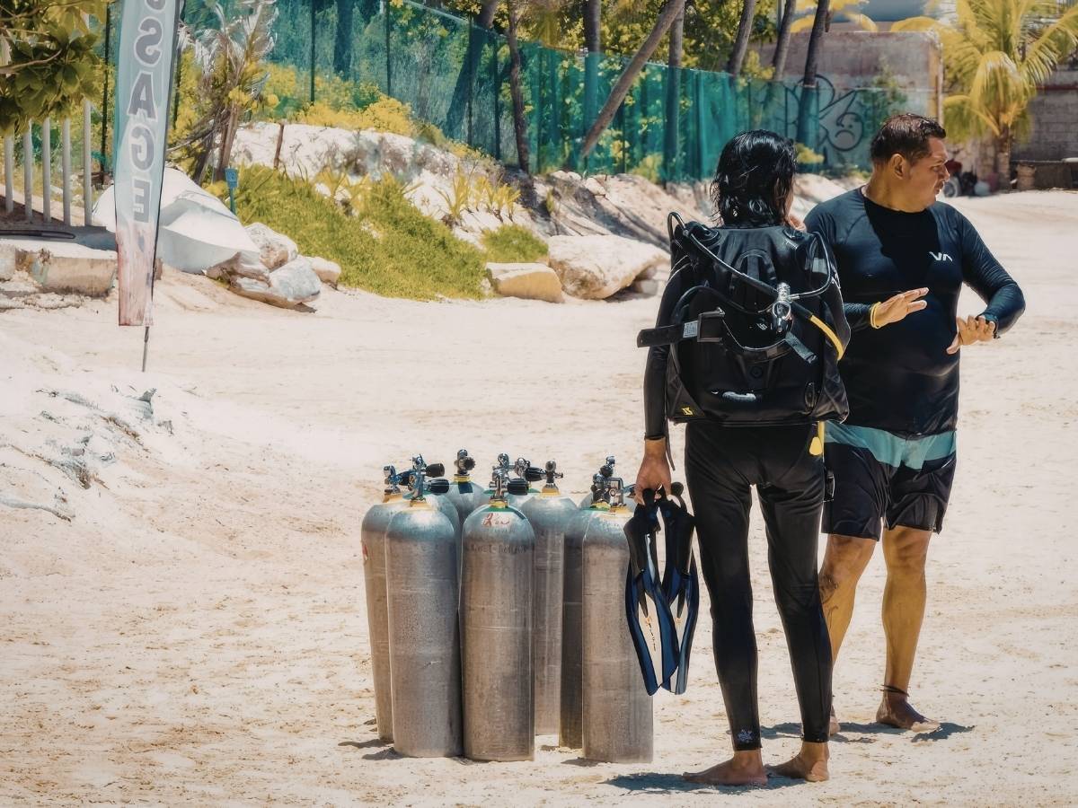 Two people in diving gear standing on a sandy beach beside several air tanks